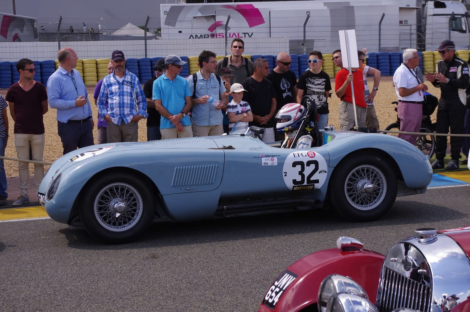A pale blue 1951 Jaguar C-Type with racing number 32 in a classic race paddock, other cars alongside