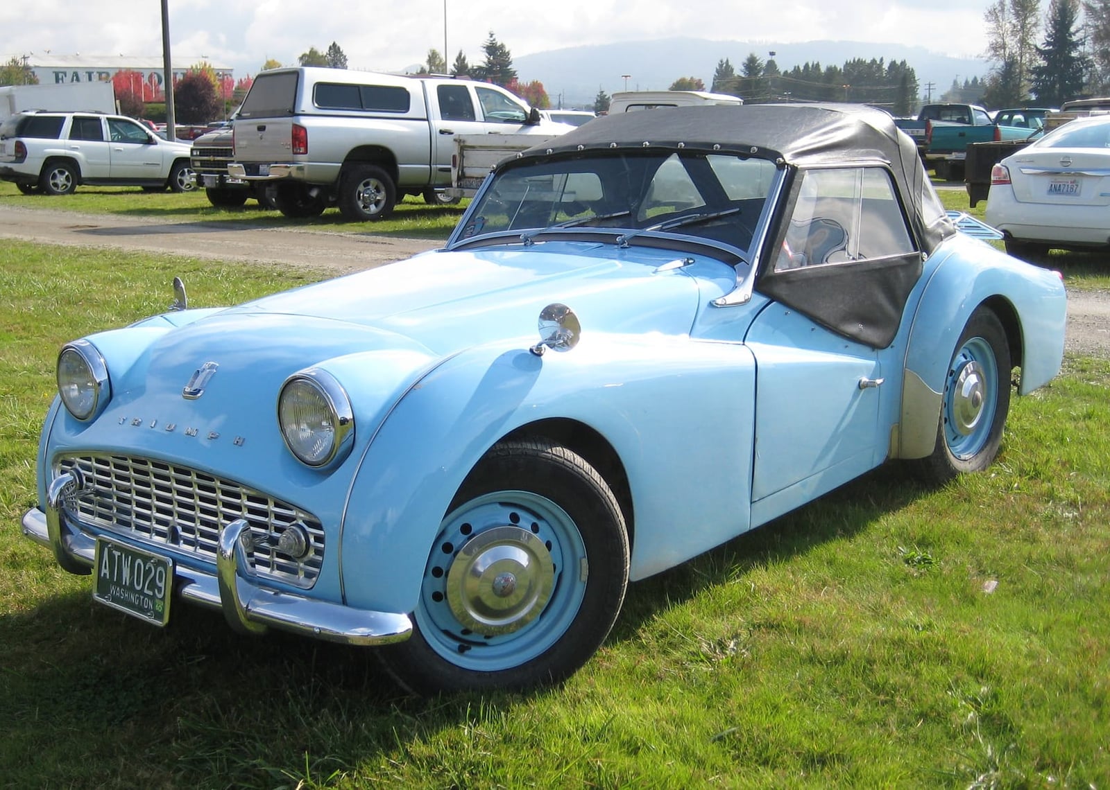 A pale blue 1960 Triumph TR3A with the hood raised and sidescreens fitted, on grass at a car meet