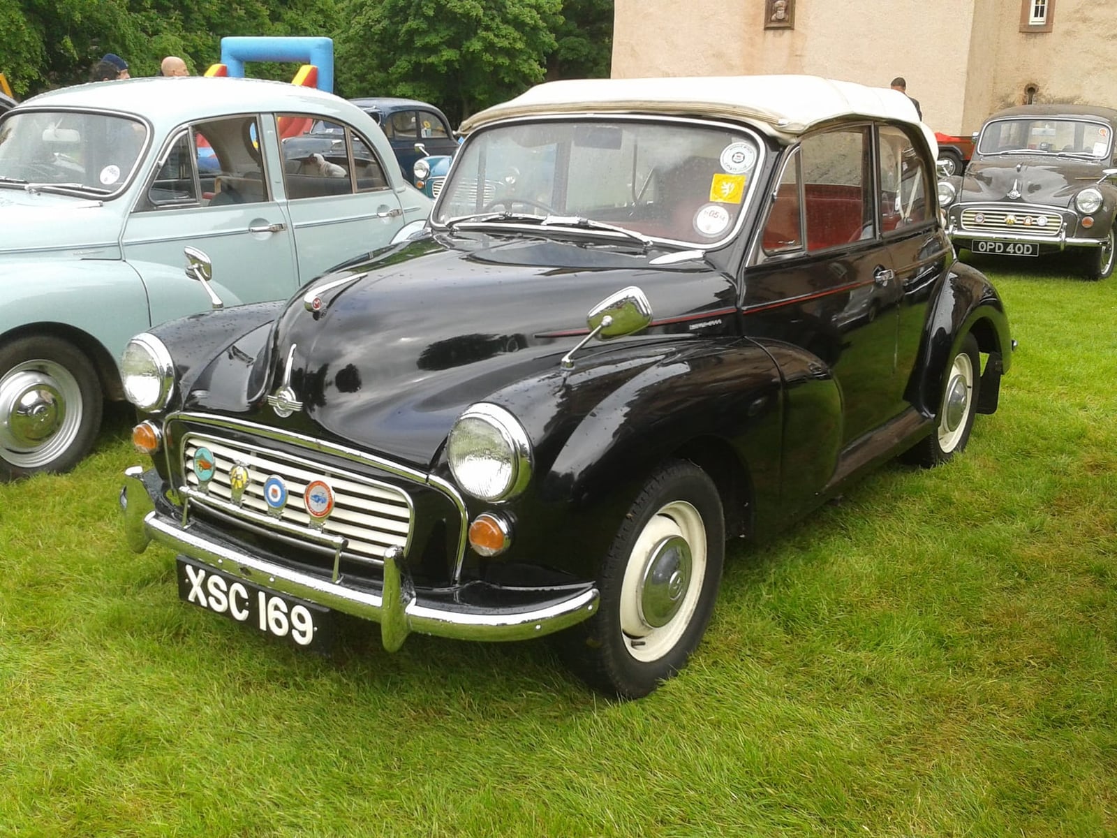 A black 1961 Morris Minor convertible with a cream hood and a UK registration, parked on grass at a show
