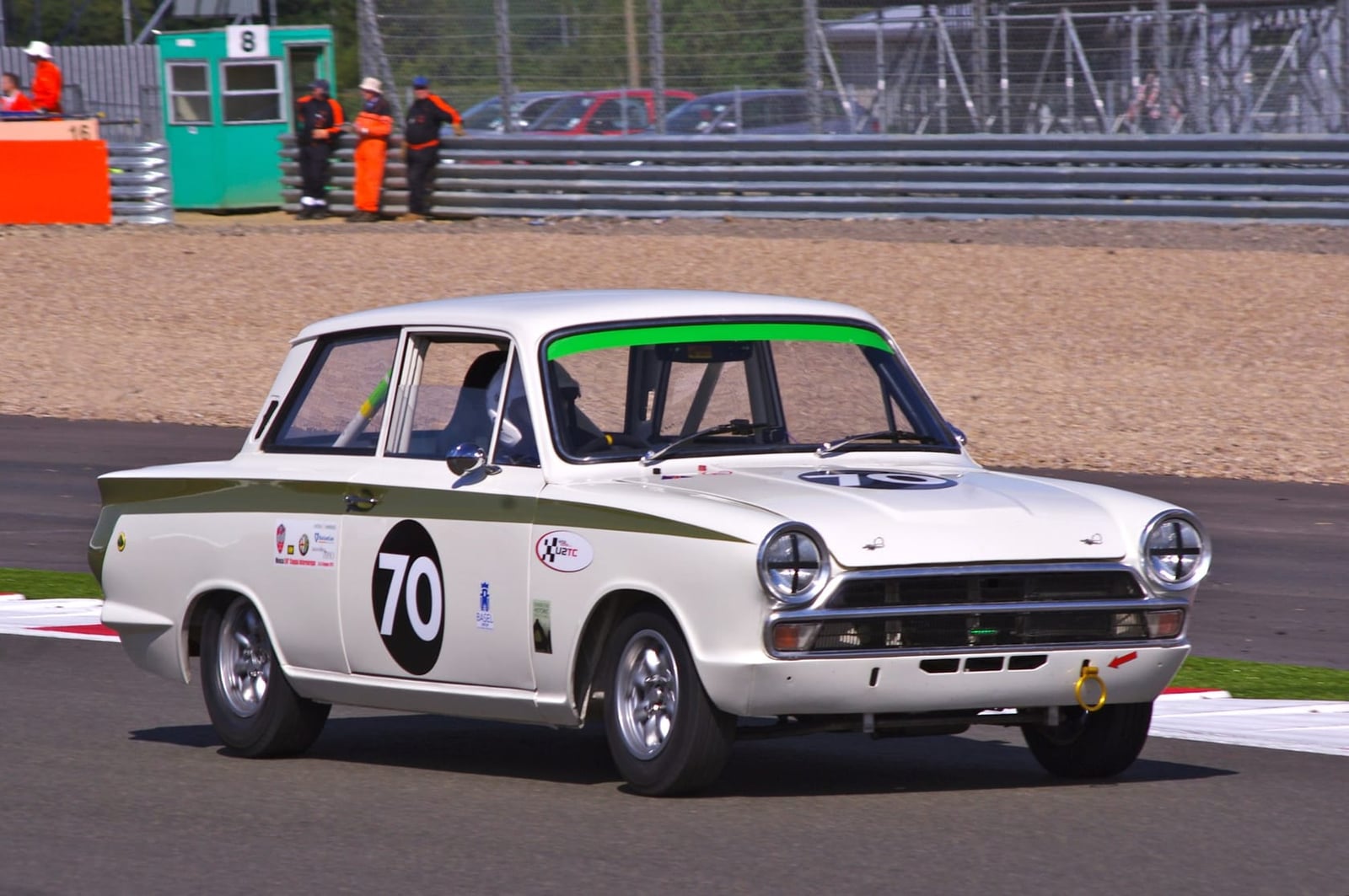 A white and green Lotus Cortina racing on a circuit, number 70, front wheel lifting