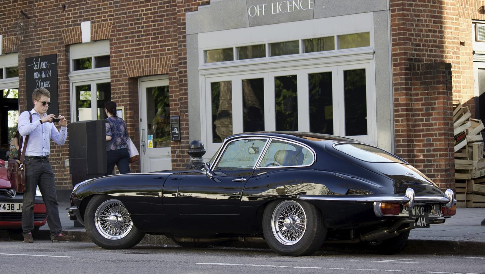 A dark Jaguar E-Type Series 2 coupe parked on a street outside a shop, side view