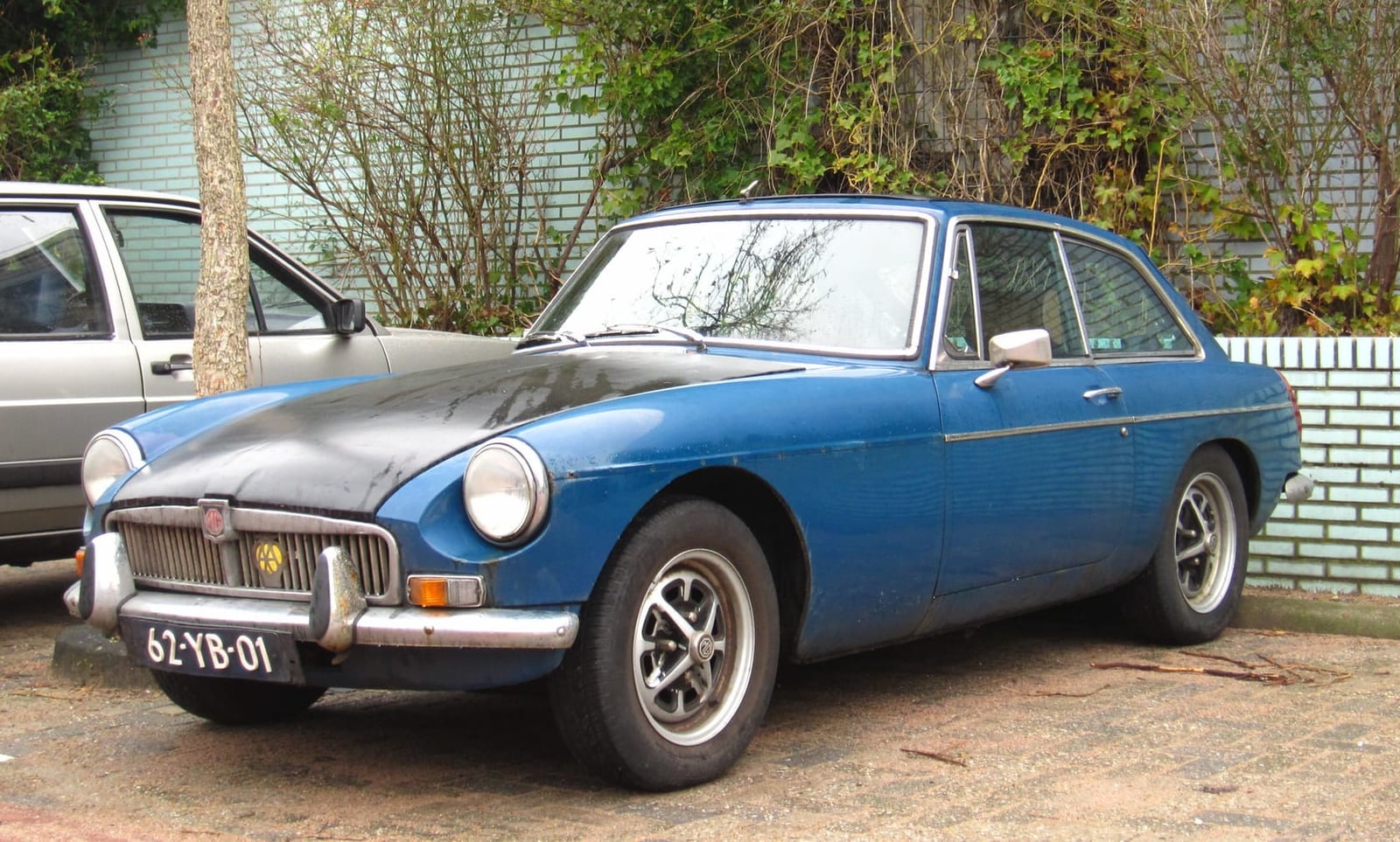A faded blue 1974 MGB GT with dull paint and a bent bumper parked on a street