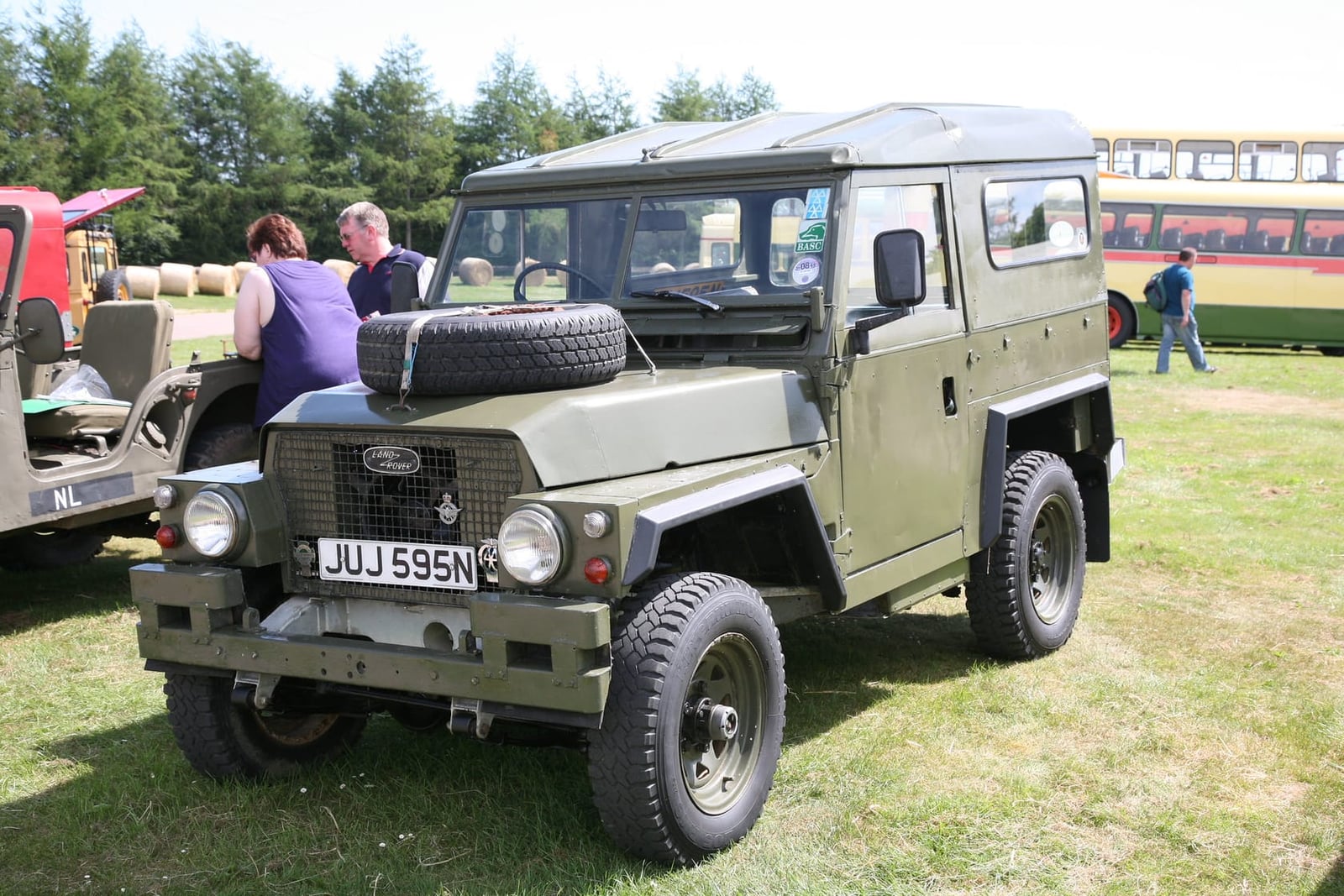 A green 1975 Land Rover Lightweight with a spare wheel on the bonnet and doors removed, on grass at a military vehicle show, UK registration JUJ 595N