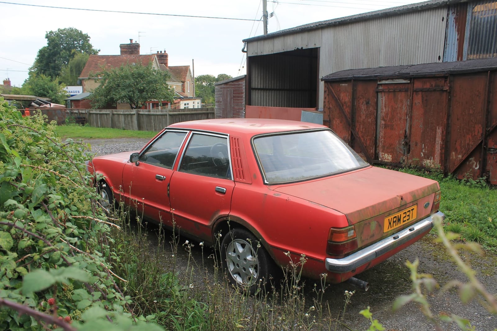 A faded red Ford Cortina Mk4 four-door saloon, rear three-quarter view, parked in long grass beside a barn