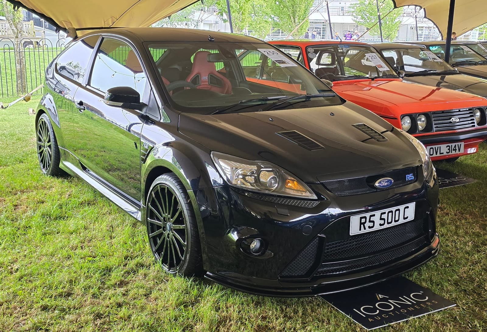 A matte black 2010 Ford Focus RS500 with a UK registration on a show field beside a Mk1 Escort