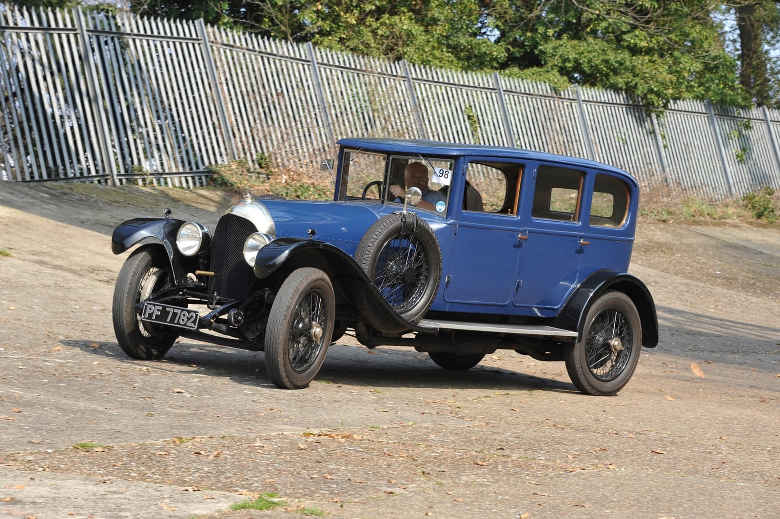 A blue 1920s Bentley 3 Litre saloon parked on the surviving concrete banking at Brooklands, palisade fence and trees behind