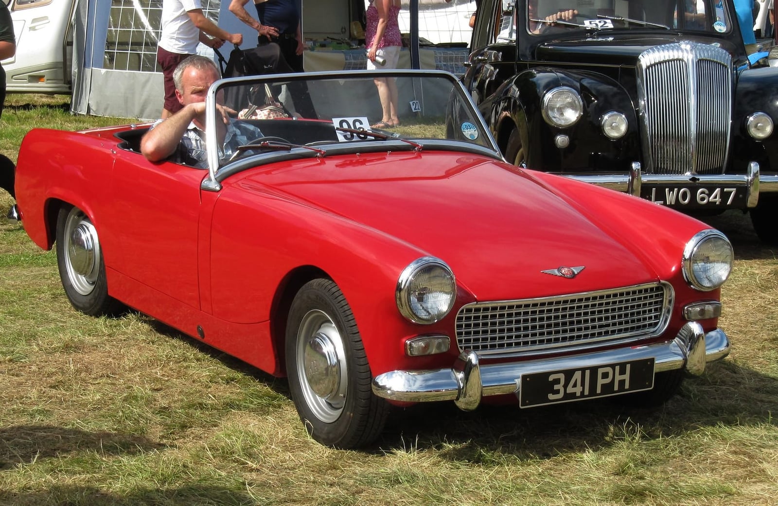 A red Austin-Healey Sprite Mk II with a UK registration at a rally, a 1950s saloon behind it