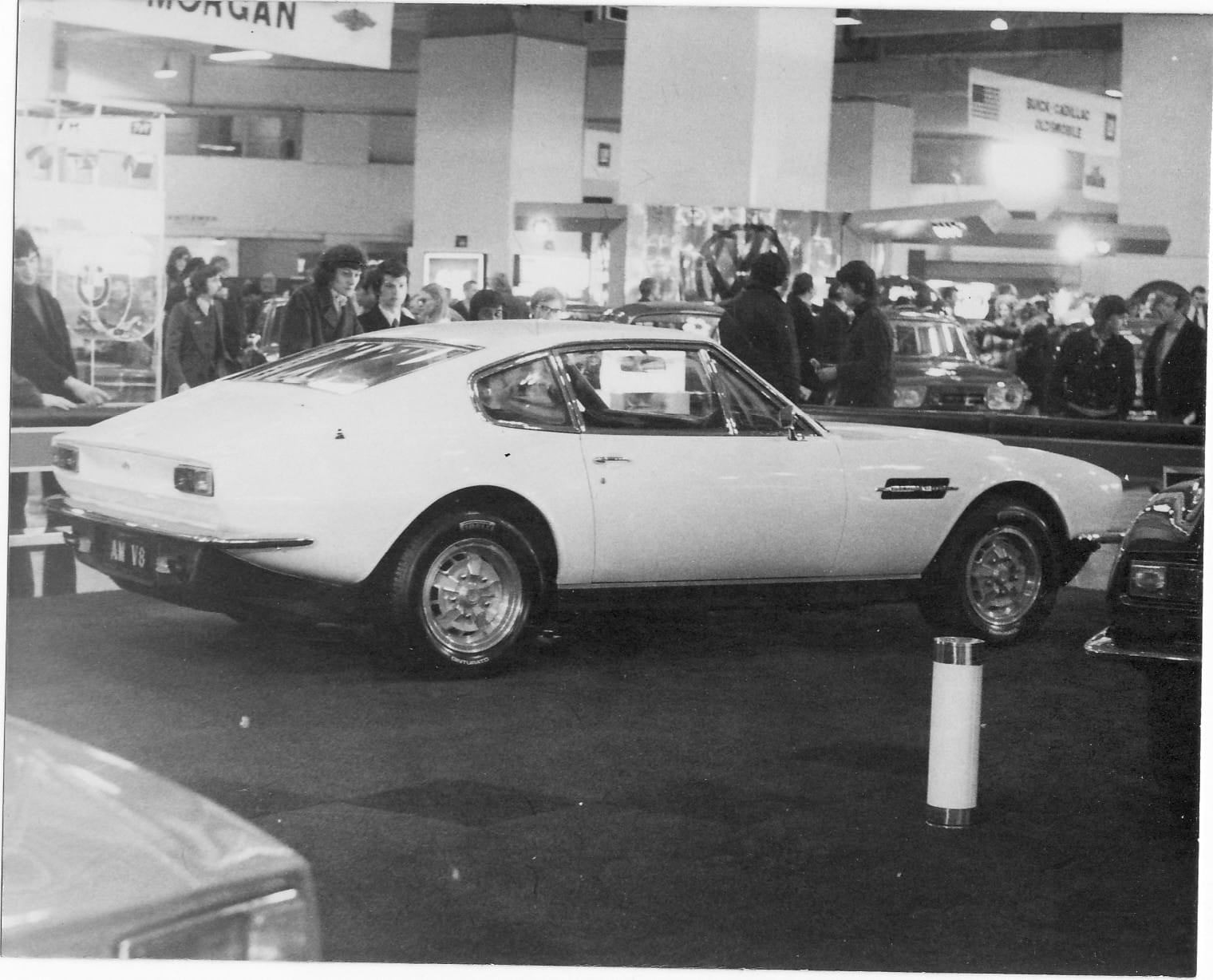 A black and white period photograph of the Aston Martin DBS V8 on a motor show stand