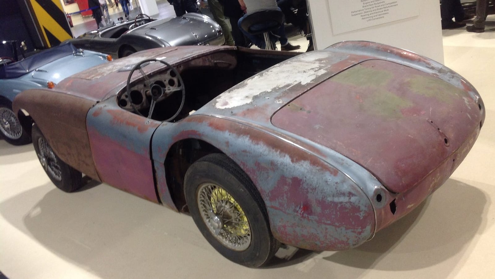 An unrestored Austin-Healey 100 bodyshell in faded red paint with wire wheels, displayed at a show