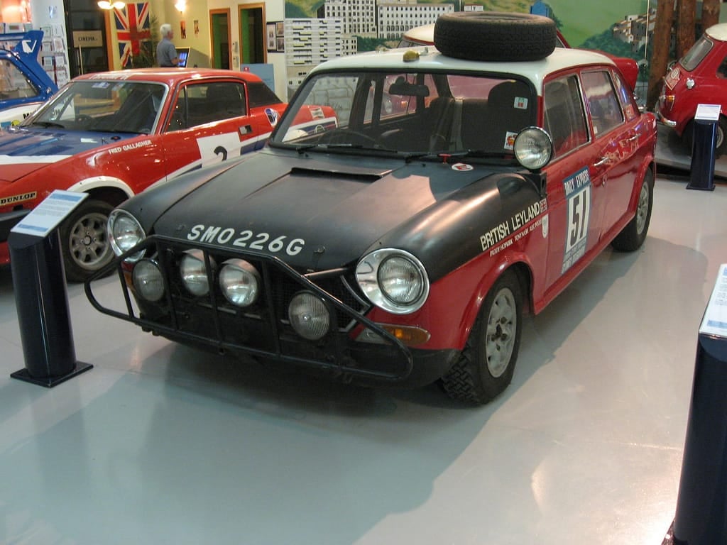 A red and black British Leyland Austin 1800 rally car with roof-mounted spare wheel and lamp bar on museum display, UK registration SMO 226G