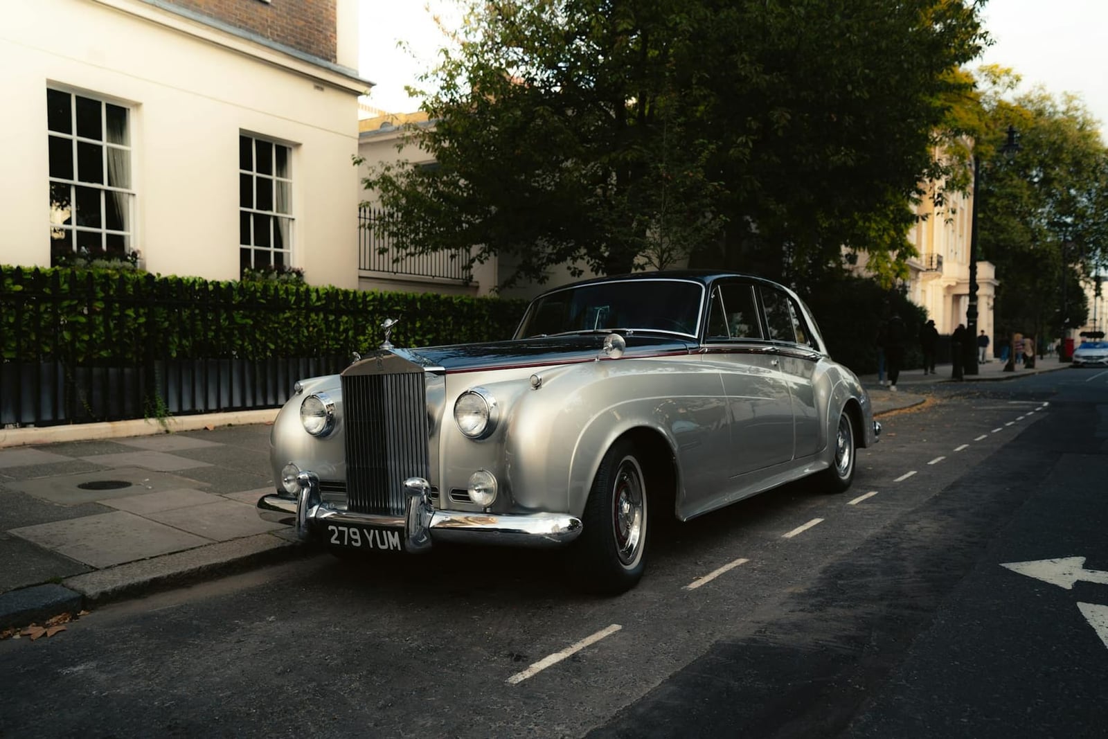 A silver Rolls-Royce Silver Cloud parked on a London street of pale stucco houses in low evening sun