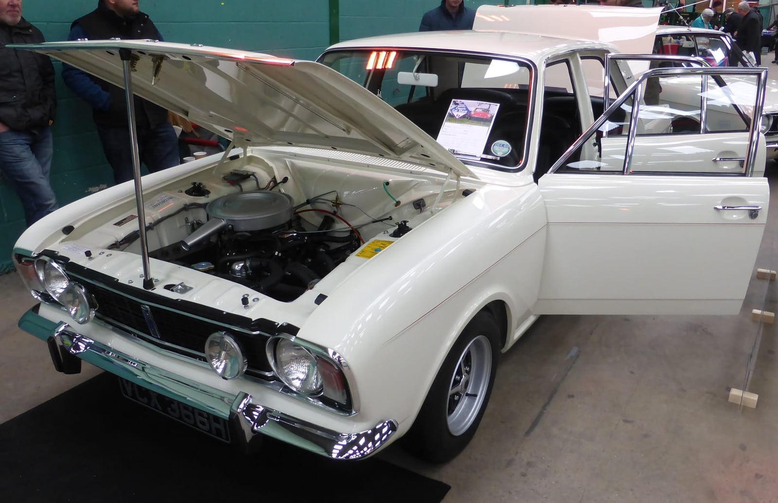 A white 1969 Ford Cortina 1600E with its bonnet raised at an indoor show