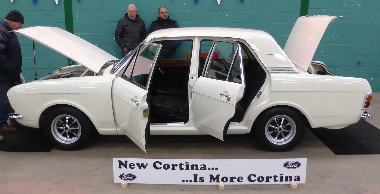 A white 1969 Ford Cortina 1600E displayed with doors open beside a period New Cortina banner