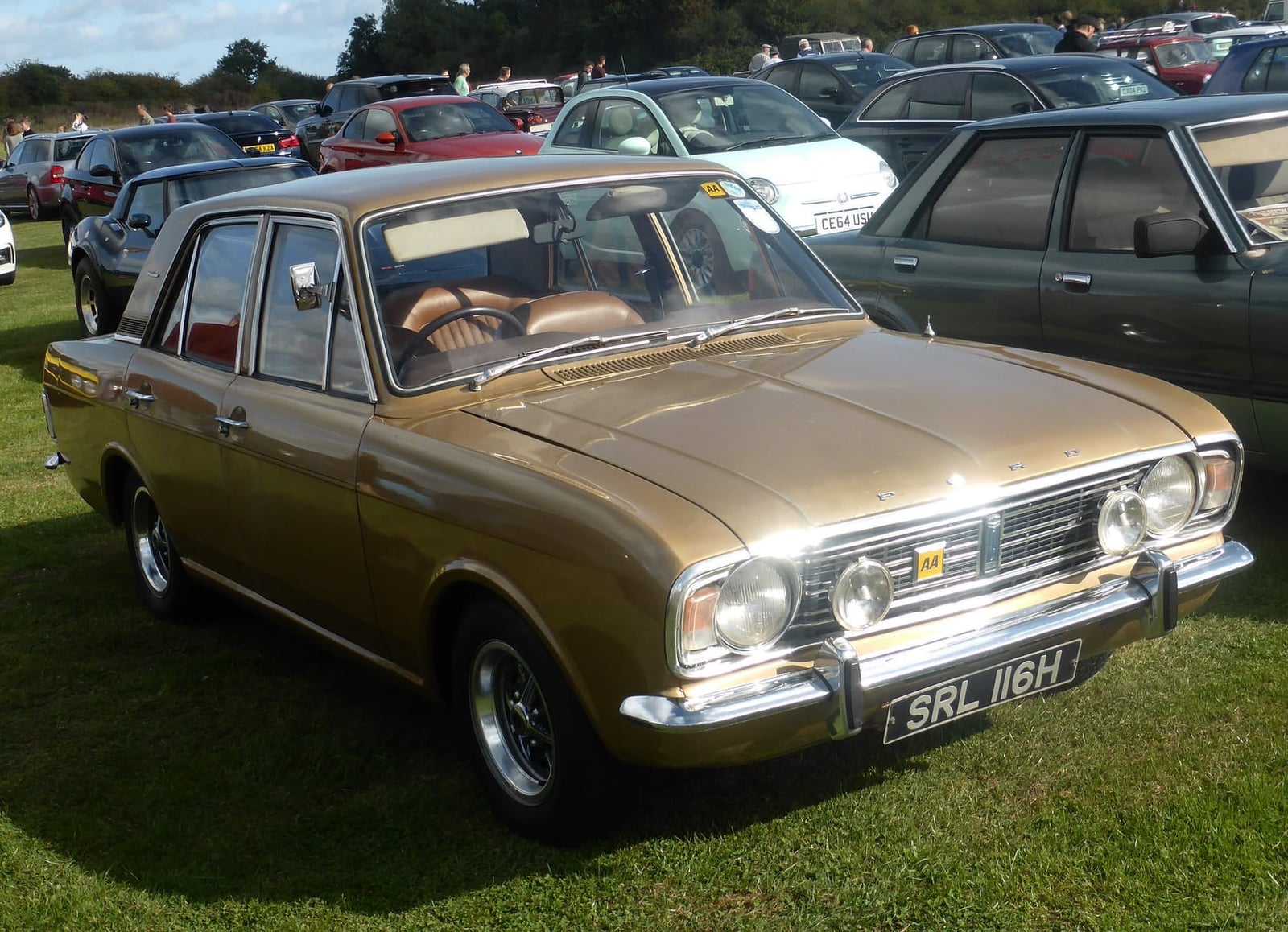 A gold 1969 Ford Cortina 1600E on grass at a classic car show, front three-quarter view, UK registration SRL 116H