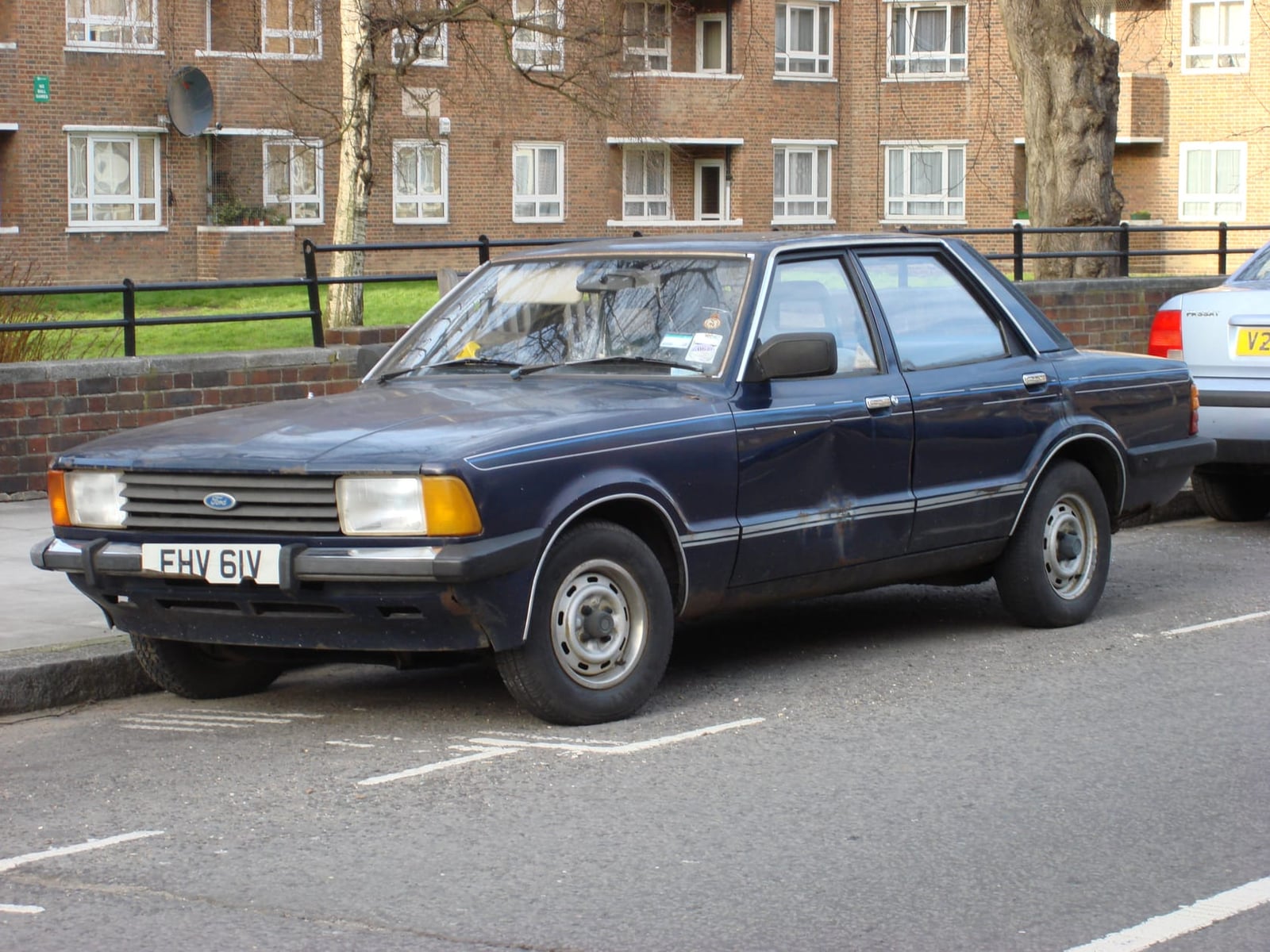 A dark blue Ford Cortina Mk4 four-door saloon parked on a British street in front of brick flats, front three-quarter view