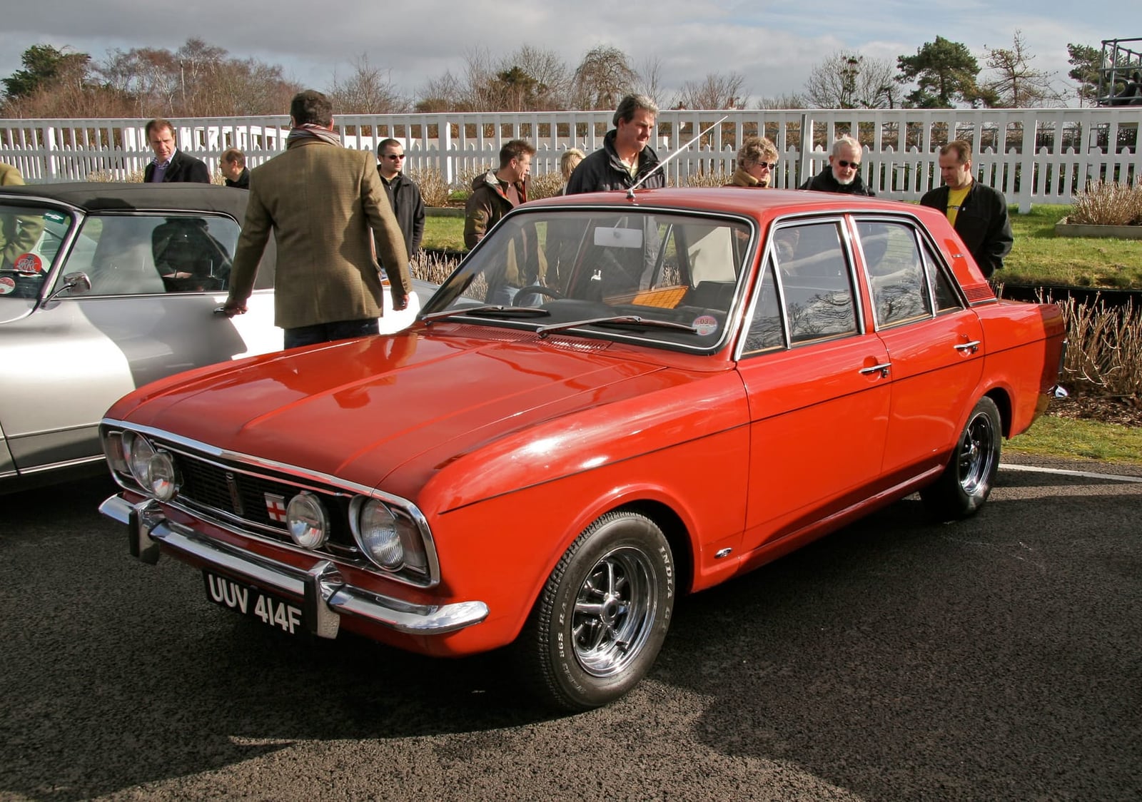 A red Ford Cortina Mk2 1600E at a race circuit paddock, front three-quarter view, UK registration UUV 414F