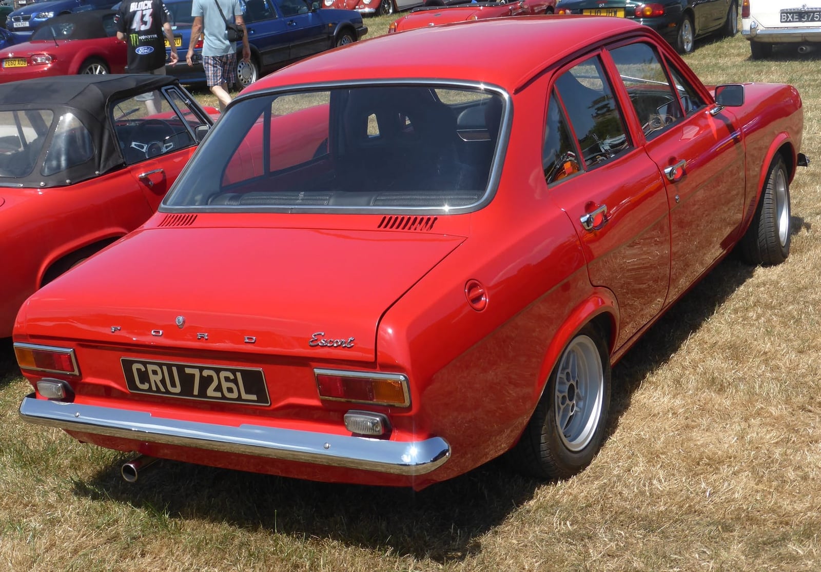 A red 1972 Ford Escort Mk1 four-door on grass at a classic car show, rear three-quarter view, UK registration CRU 726L
