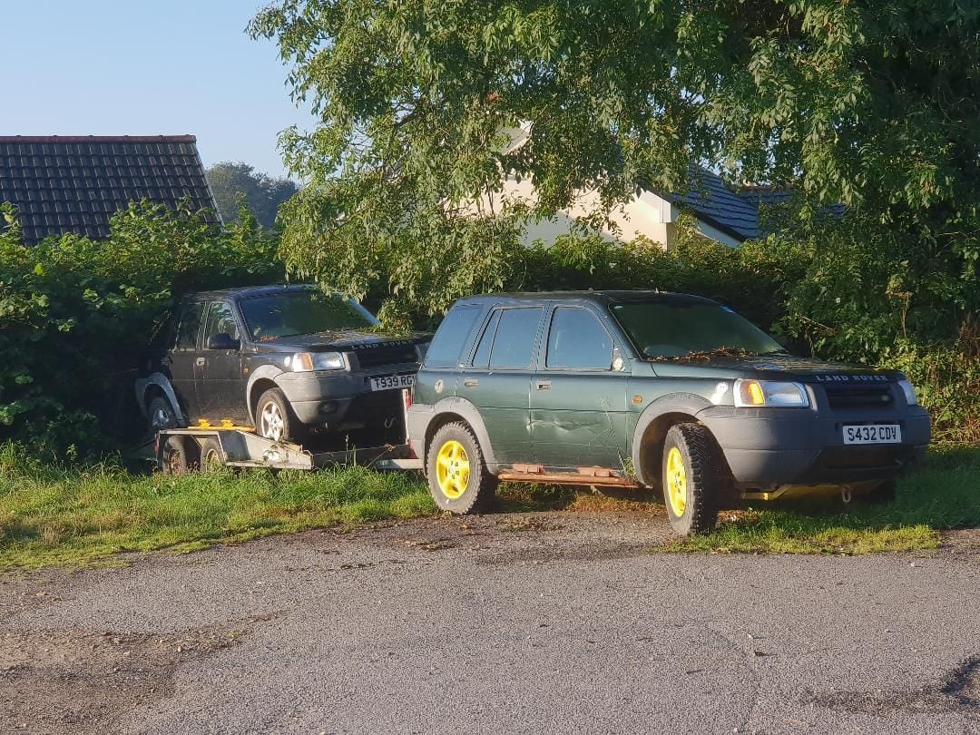 A green Land Rover Freelander 1 on a trailer being towed through a field by a Freelander 2