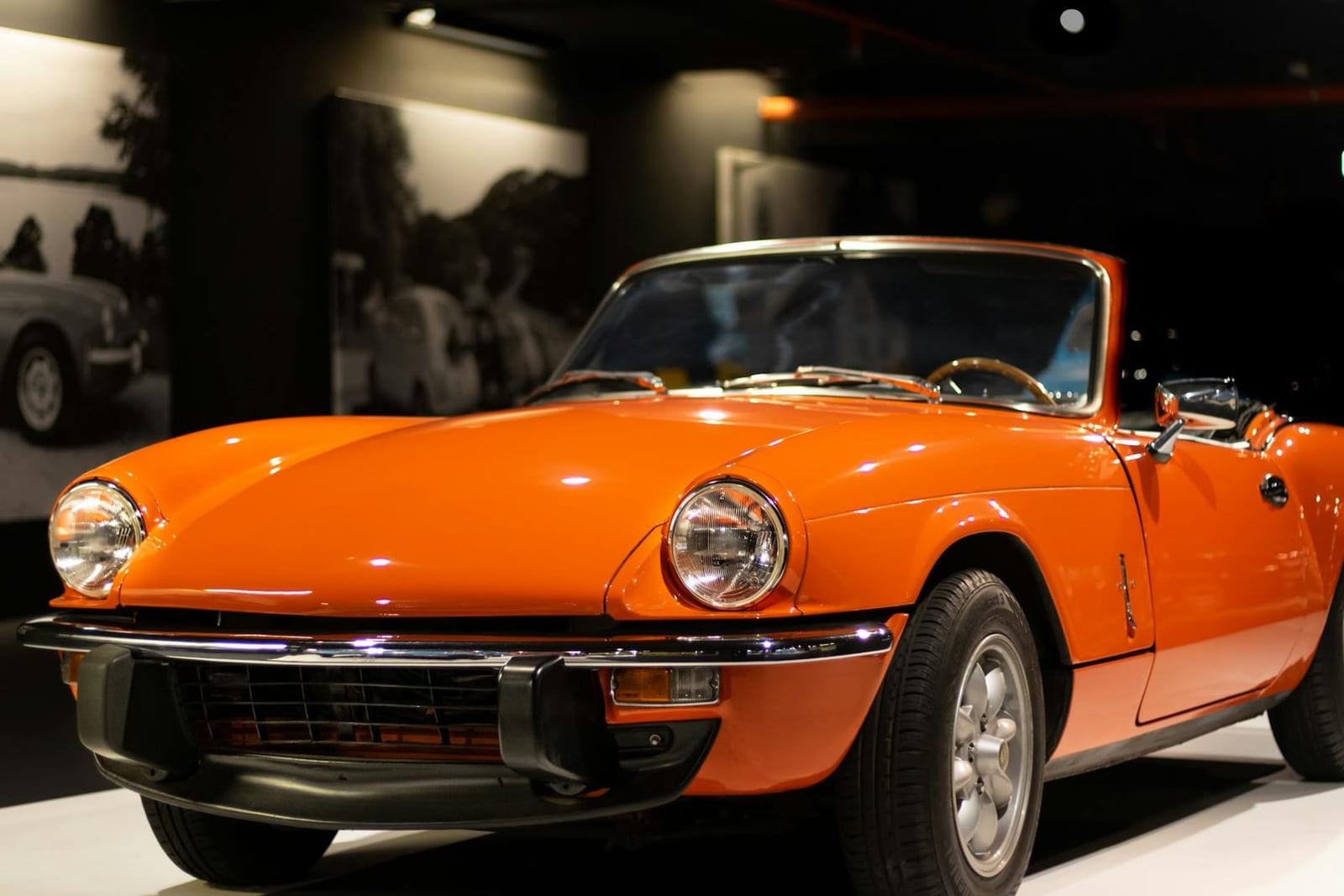 An orange Triumph Spitfire roadster, front three-quarter view, on display under exhibition lighting