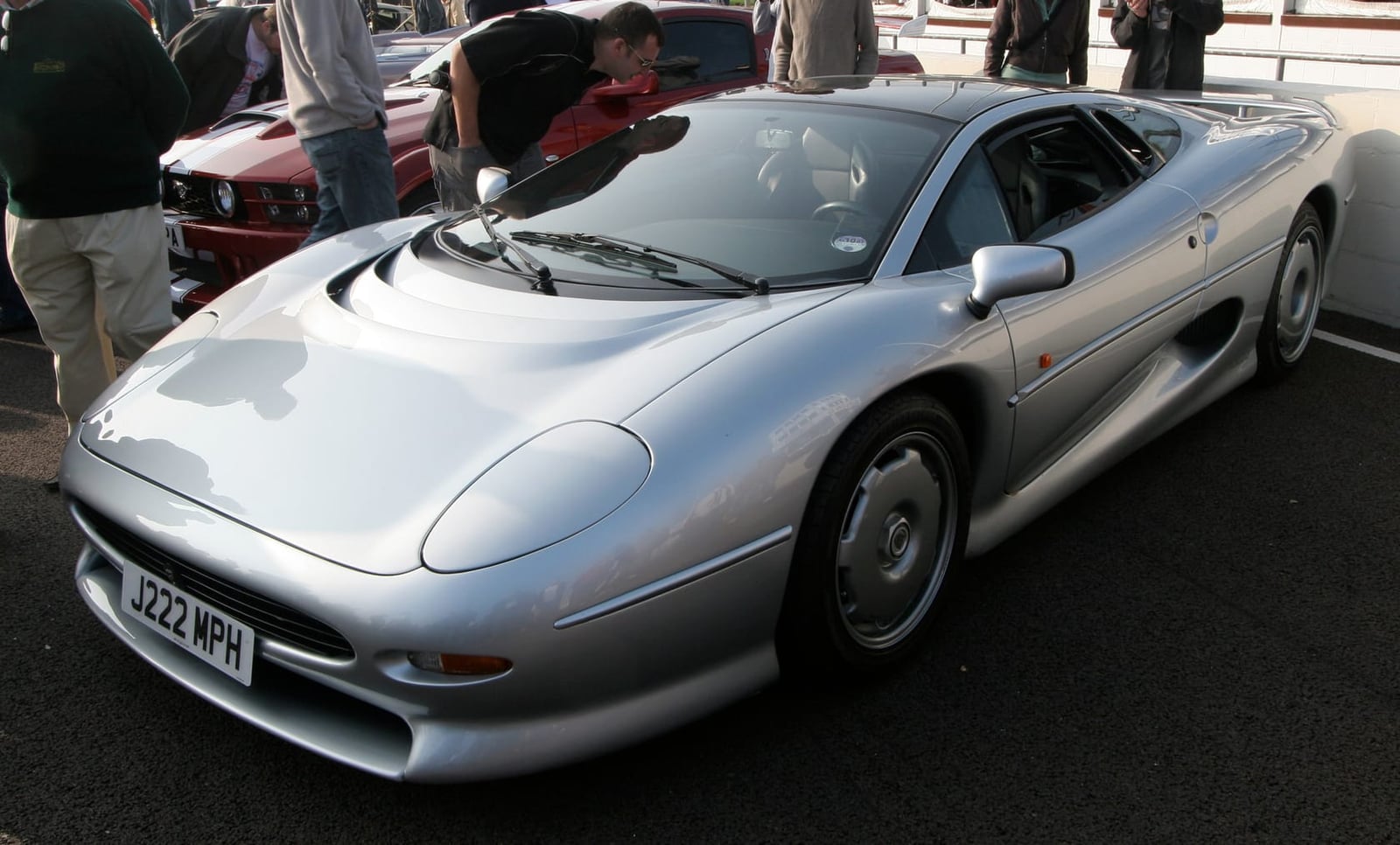 A silver Jaguar XJ220 supercar with onlookers leaning in to look, at a Goodwood Breakfast Club meet