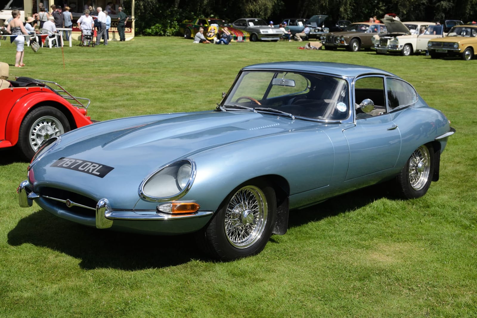 A silver-blue 1962 Jaguar E-Type Series 1 coupe on grass at a classic car gathering, front three-quarter view, UK registration 157 URL