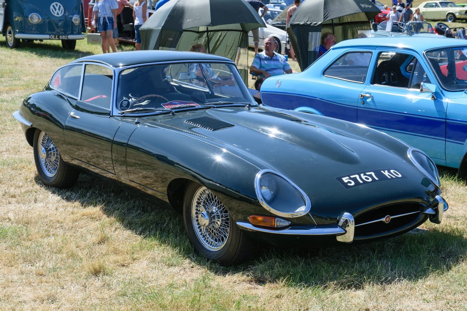 A dark blue 1964 Jaguar E-Type Series 1 coupe on grass at a classic car show, front three-quarter view, UK registration 576T KO