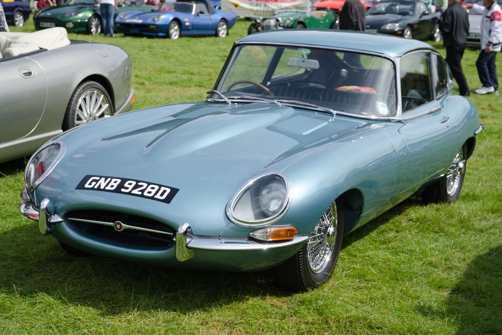 A pale blue 1966 Jaguar E-Type Series 1 fixed-head coupe on grass at a classic car show, front three-quarter view, UK registration GNB 928D