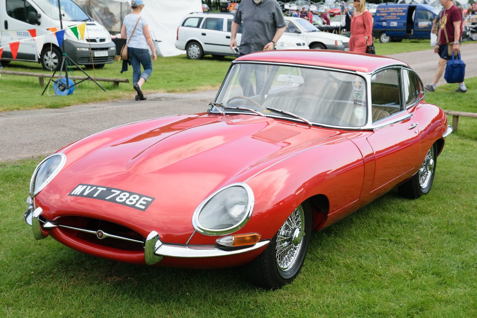 A red 1967 Jaguar E-Type Series 1 coupe on grass at a classic car show, front three-quarter view, UK registration WVT 788E