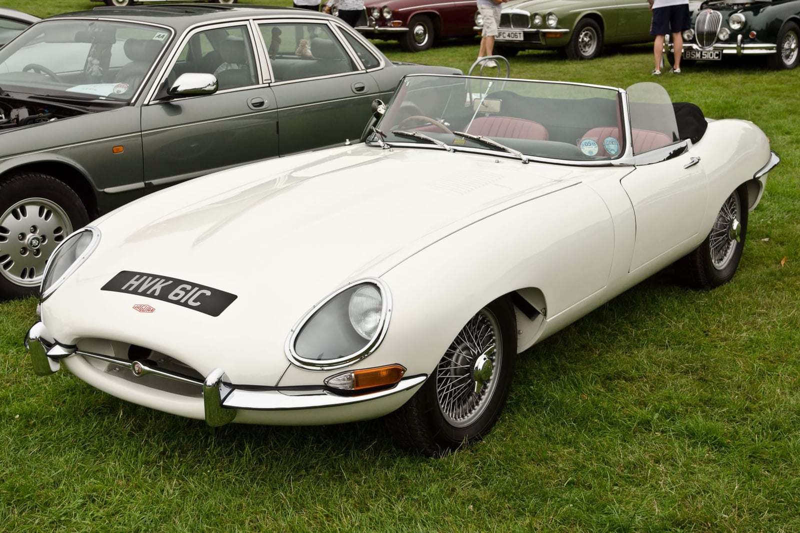 A white 1965 Jaguar E-Type Series 1 roadster on grass at a classic car show, front three-quarter view, UK registration HVK 61C