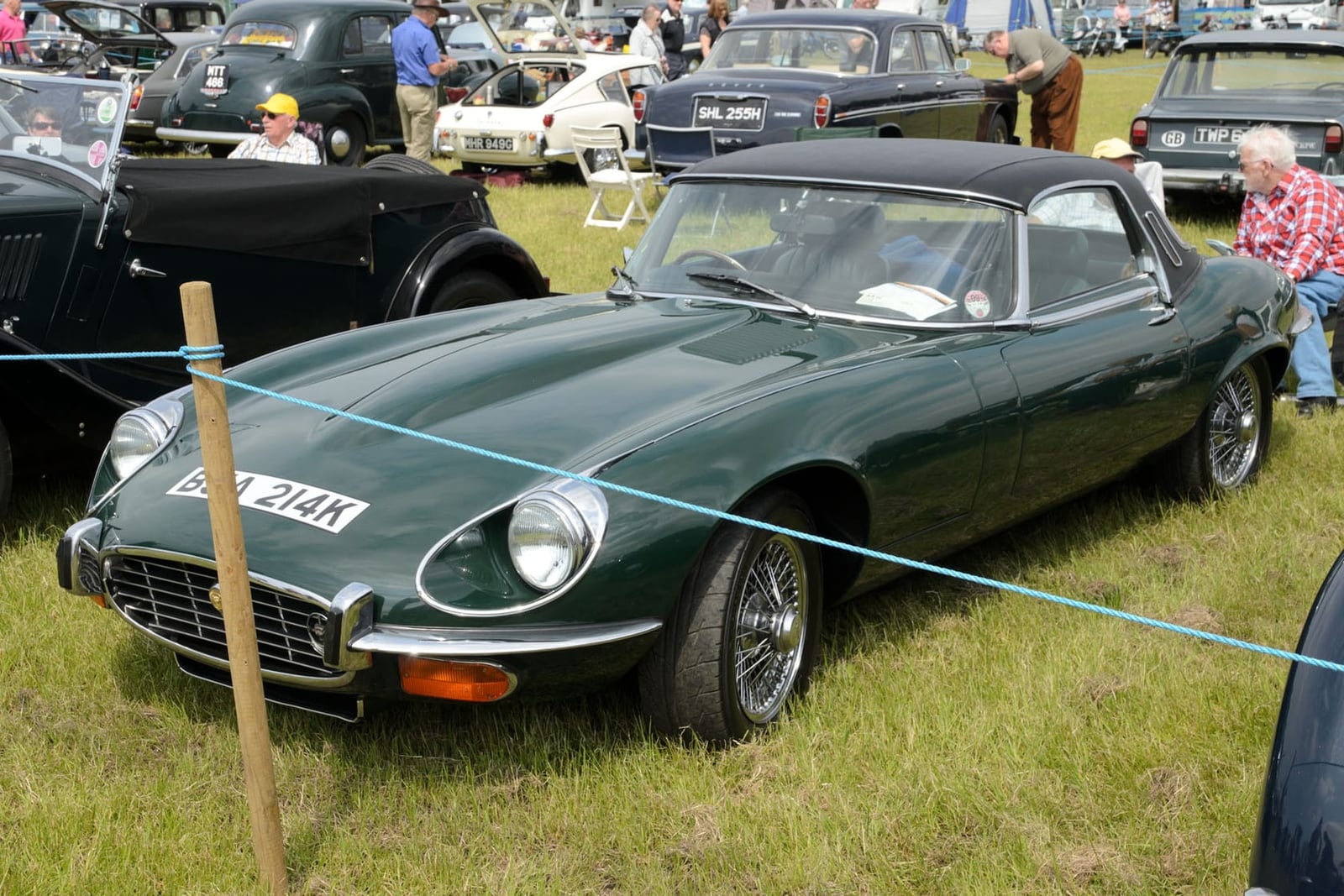 A dark green 1971 Jaguar E-Type Series 3 roadster on grass at a classic car show, front three-quarter view, UK registration BBD 214K