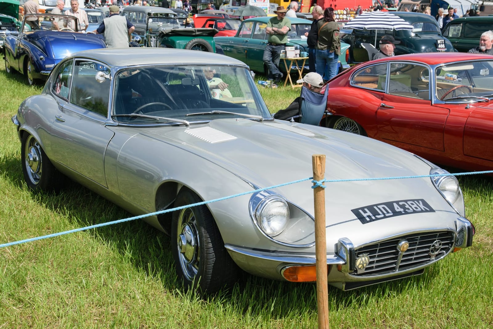 A silver 1972 Jaguar E-Type Series 3 V12 coupe on grass at a classic car show, front three-quarter view, UK registration HJD 438K
