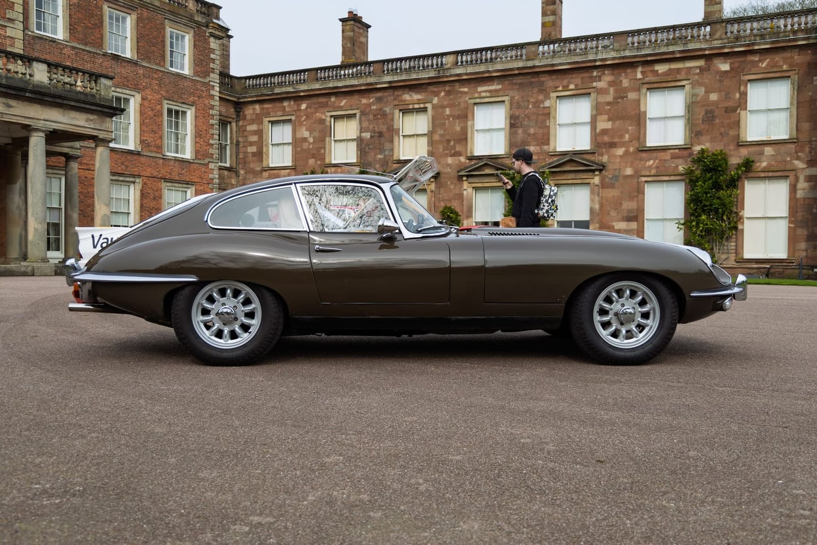A grey Jaguar E-Type fixed-head coupe in side profile, parked in front of a grand brick country house
