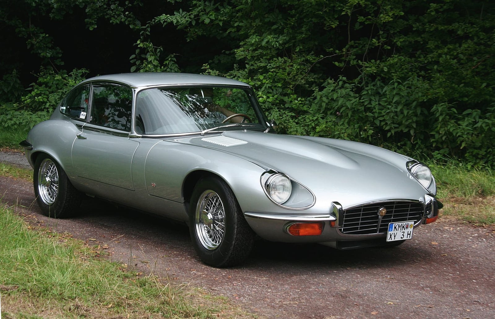 A silver Jaguar E-Type Series 3 V12 coupe parked beside trees, front three-quarter view