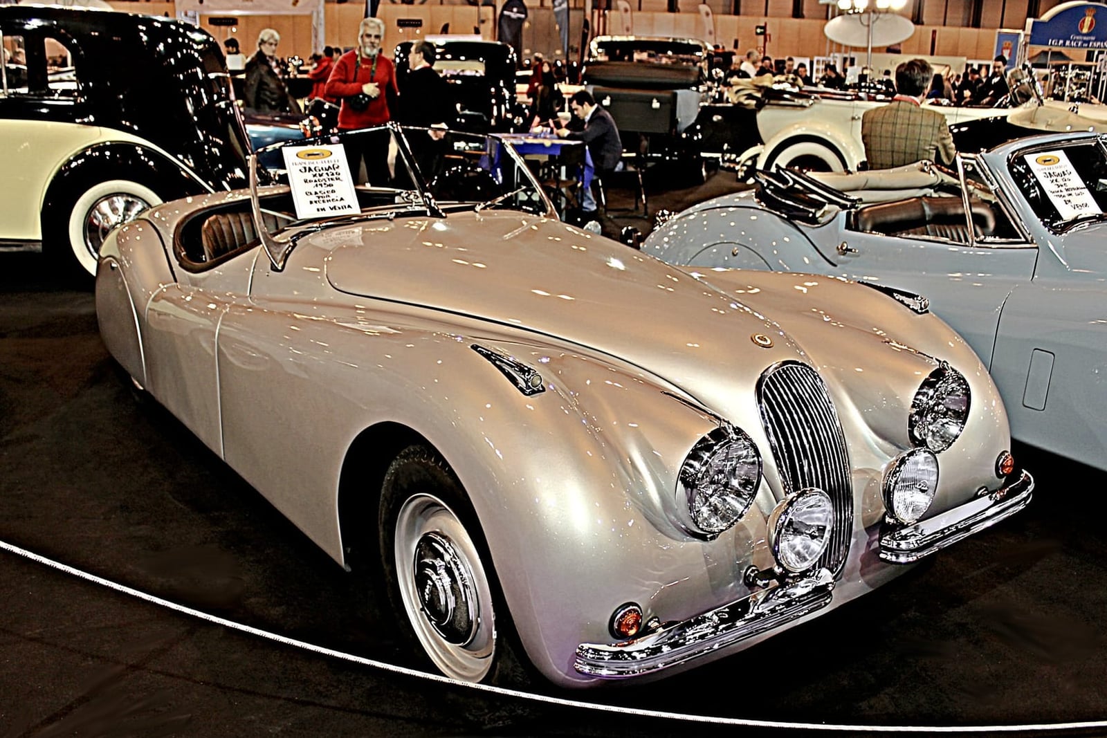 A silver 1950 Jaguar XK120 roadster displayed on a turntable at an indoor concours among other cars