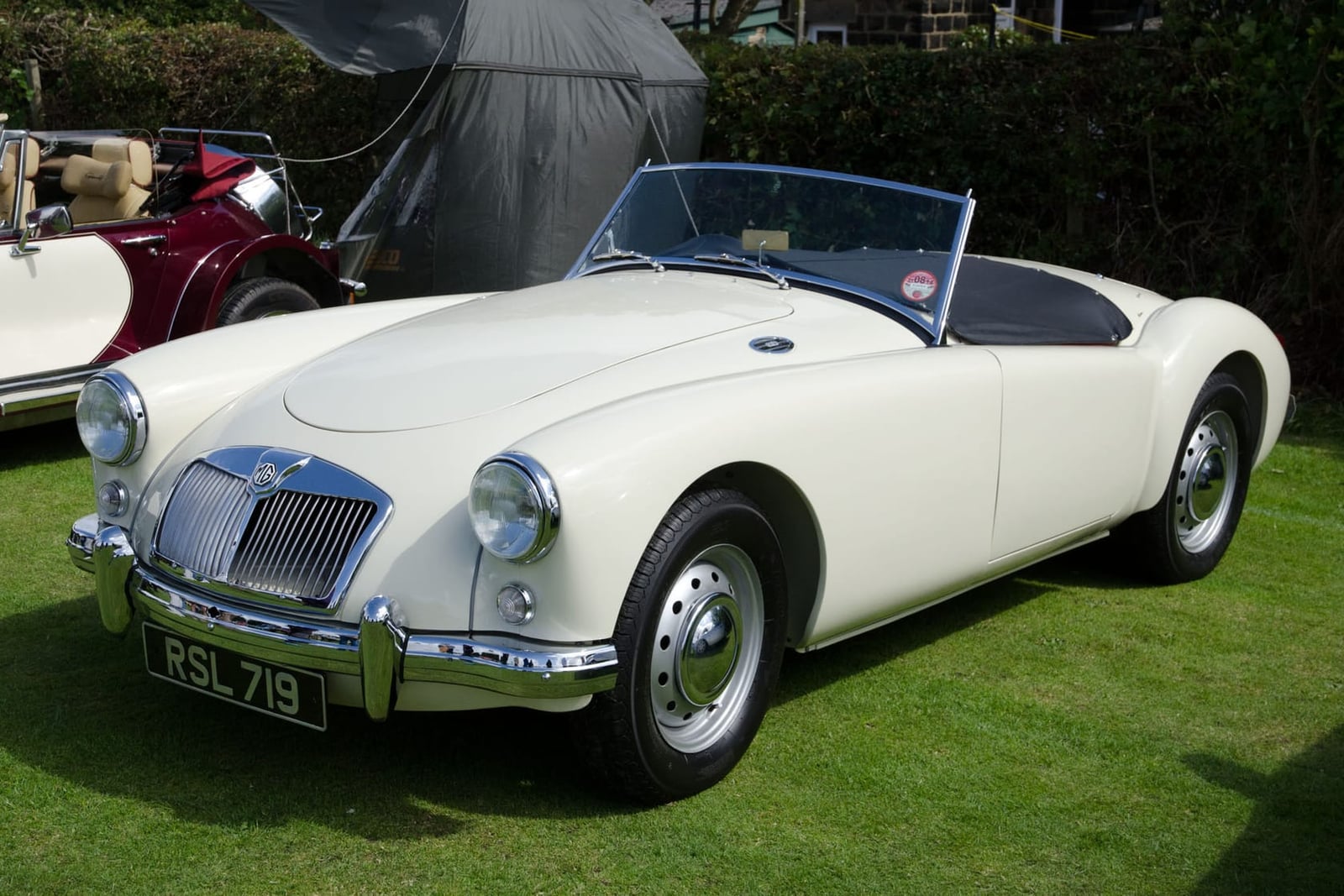 A cream 1956 MGA 1500 roadster with a UK registration parked on grass at a show