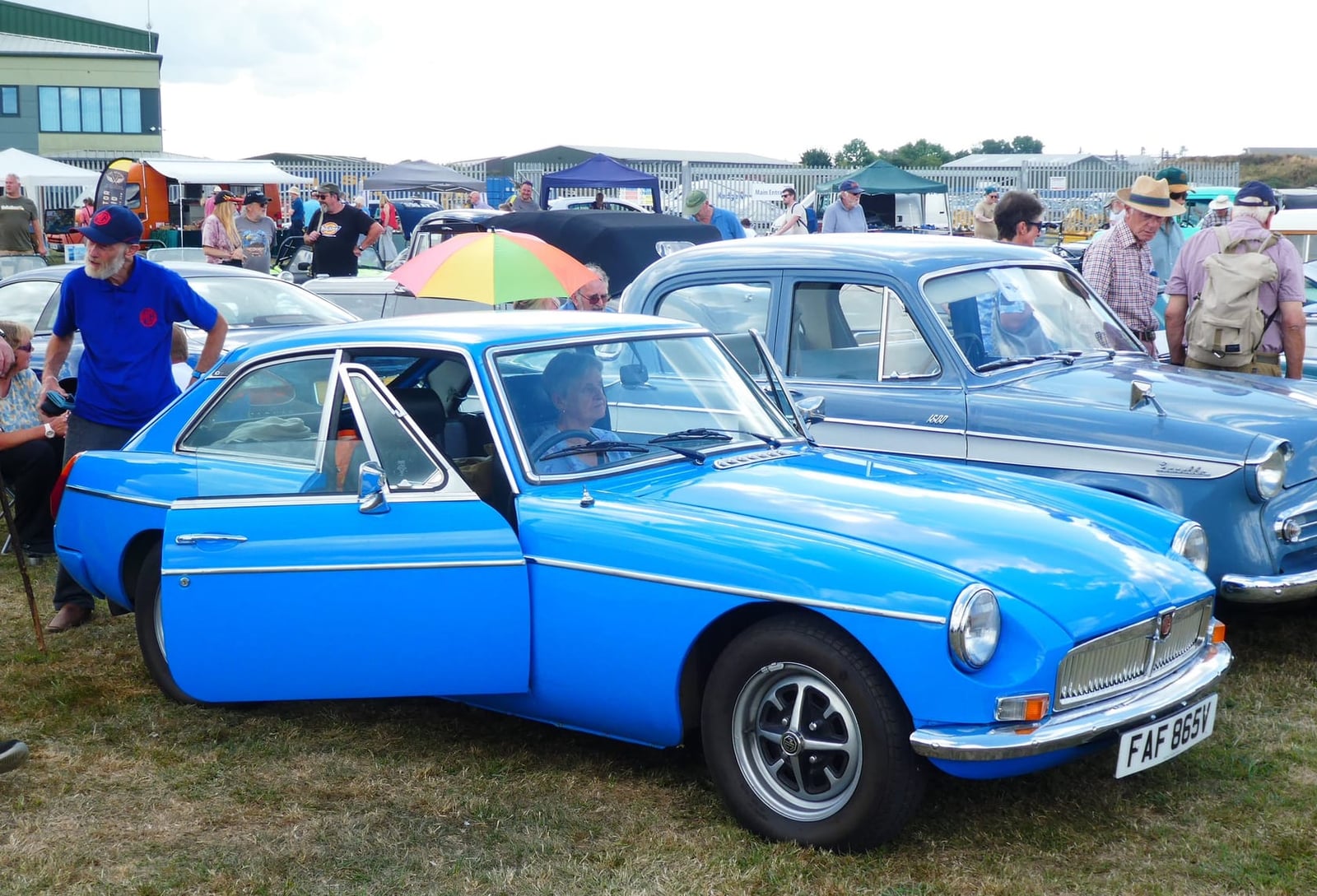 A bright blue 1980 MGB GT with black rubber bumpers and a UK registration at a busy show