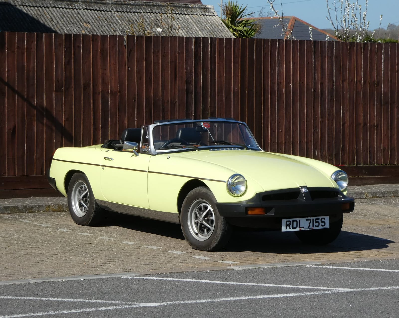 A primrose yellow MGB roadster with its hood down, registration RDL 715S, parked in front of a wooden fence in sunshine