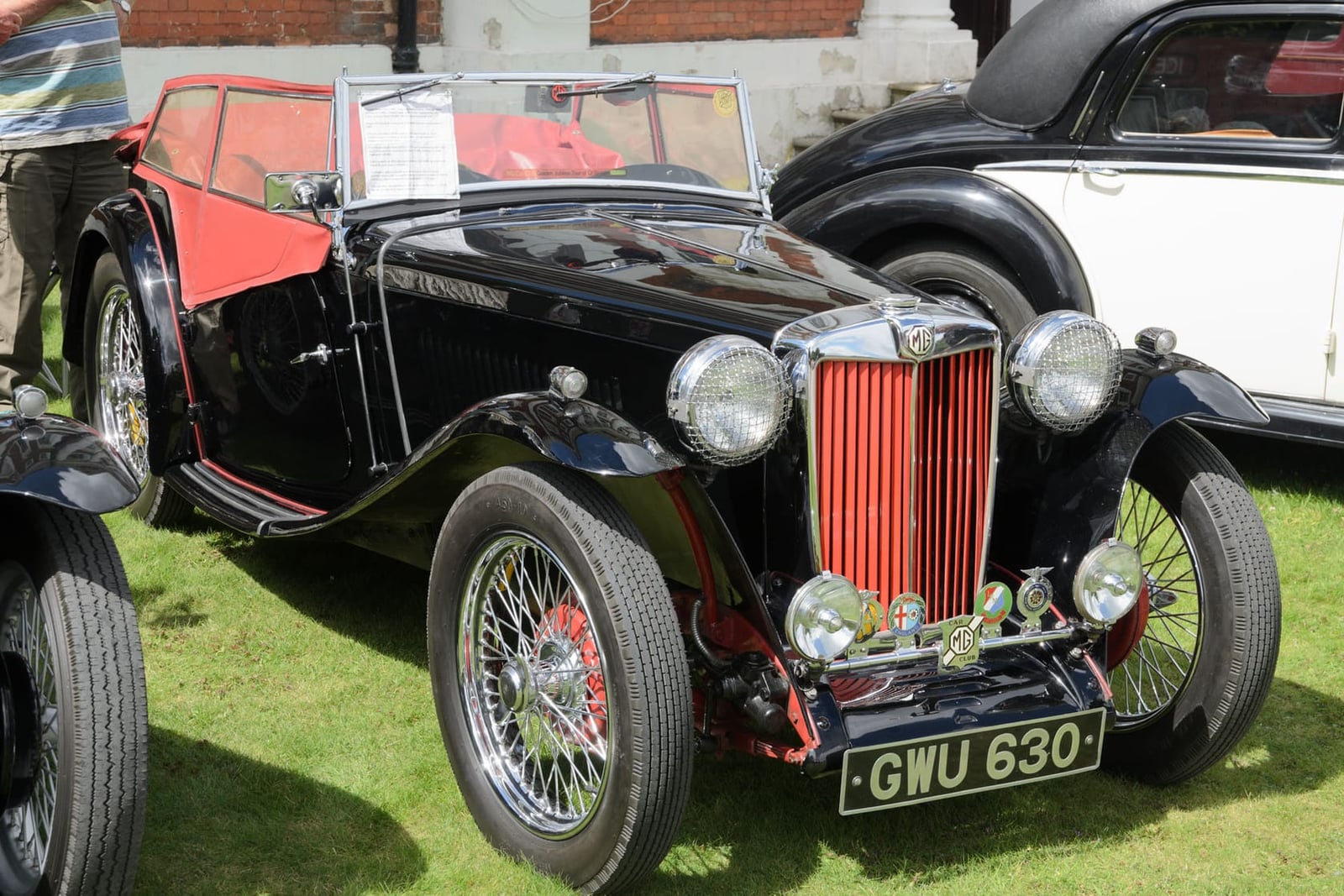 A black 1947 MG TC with a UK registration and wire wheels parked on grass beside a pre-war saloon