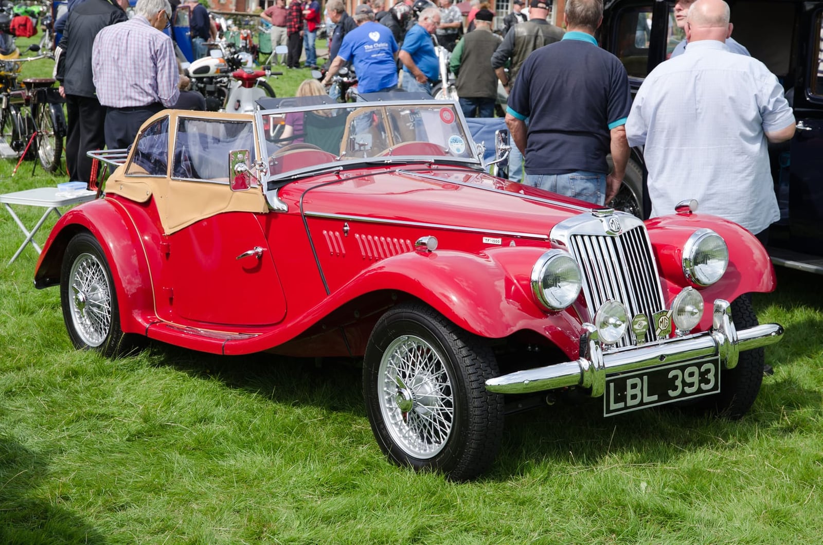A red 1955 MG TF 1500 with a UK registration on a show field, other classics behind