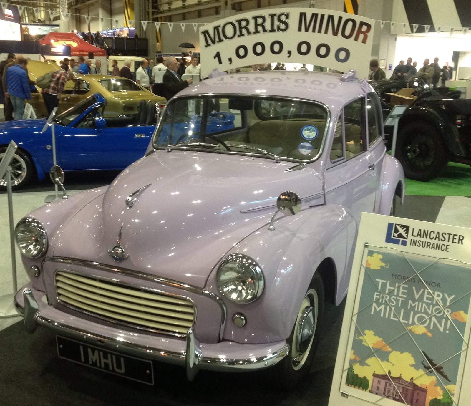 A lilac 1961 Morris Minor Million with the registration 1 MHU, displayed at an indoor show beneath a banner reading Morris Minor 1,000,000