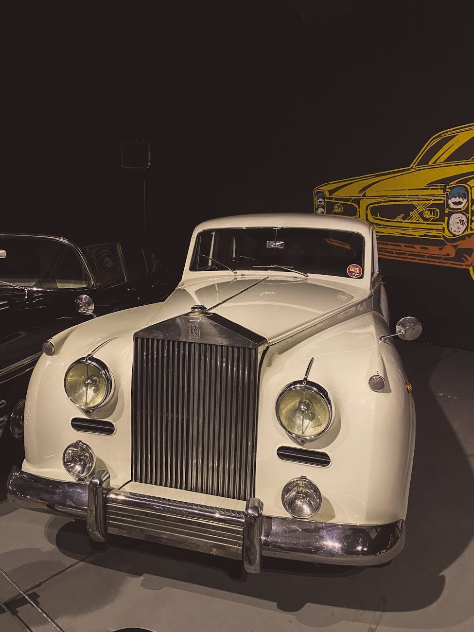 A white Rolls-Royce Silver Cloud photographed at night beside a yellow classic car