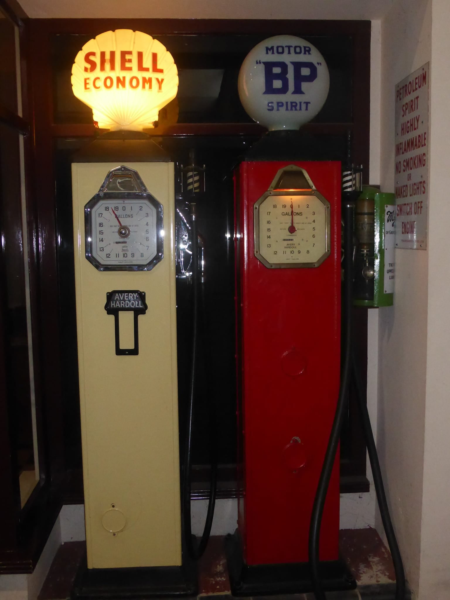 Two restored pre-war petrol pumps, a cream Shell Economy pump and a red BP Motor Spirit pump with illuminated glass globes, in a museum