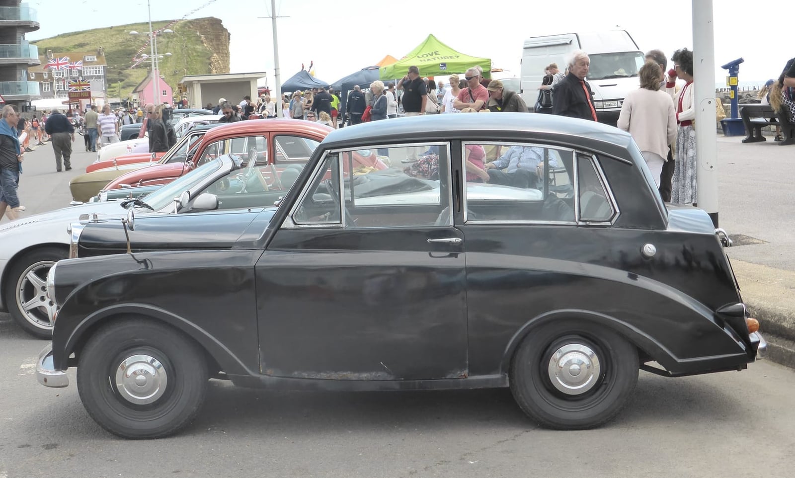A black Triumph Mayflower in side profile on a seafront promenade, showing the razor-edge roofline and upright tail