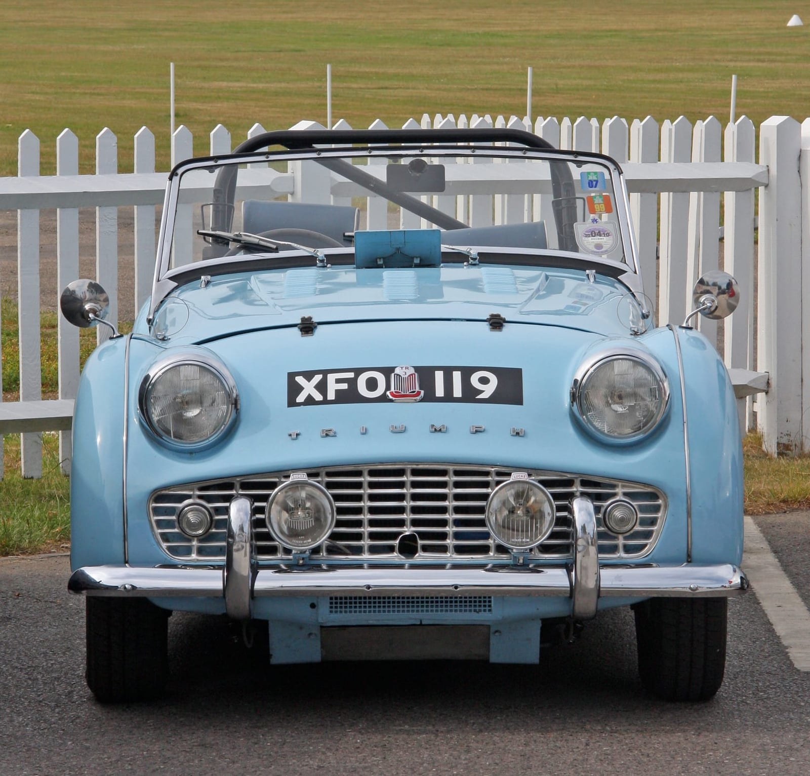A pale blue Triumph TR3A photographed head-on beside a white fence, showing the full-width grille, UK registration XFO 119