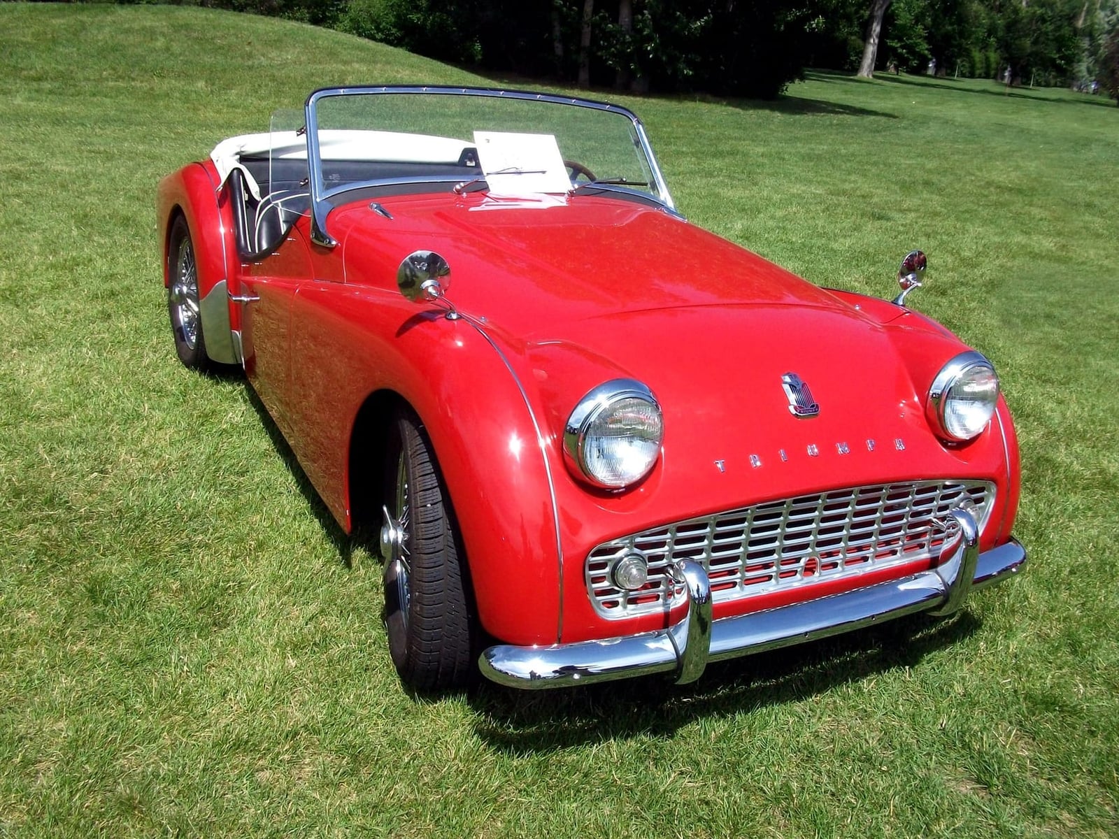 A red Triumph TR3A on grass with the hood down and windscreen upright, front three-quarter view