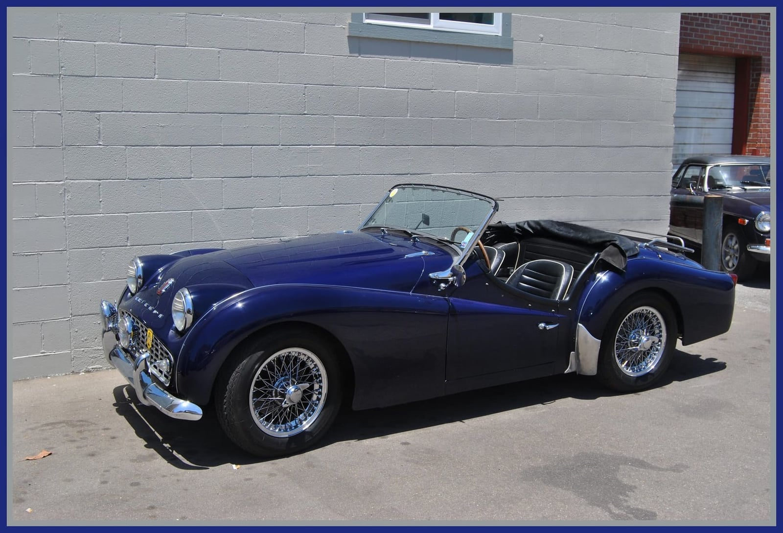A dark blue Triumph TR3A on wire wheels parked beside a wall, side view showing the cutaway doors