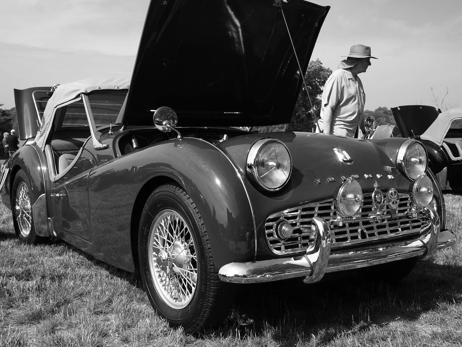 Black-and-white front three-quarter view of a Triumph TR3A with the bonnet raised at a show, showing the full-width grille