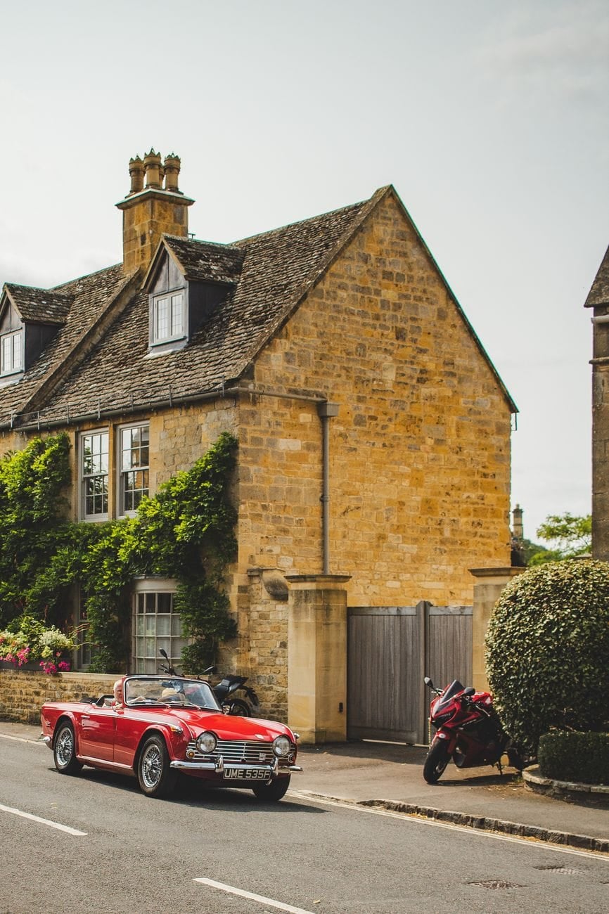 A red Triumph TR4 with the top down parked in front of a honey-stone Cotswold cottage