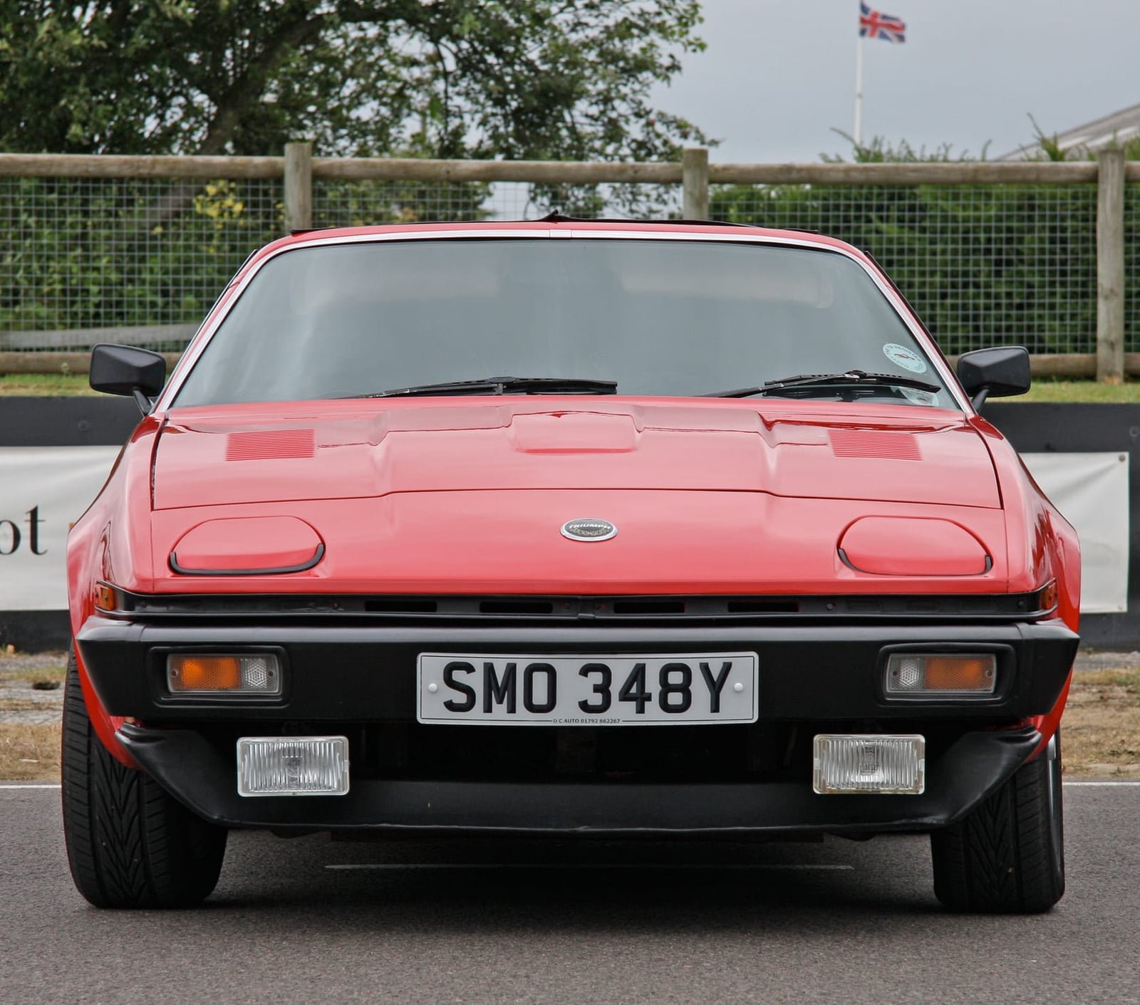 A red Triumph TR7 coupe photographed head-on, showing the pop-up headlamps closed and the black bumper, UK registration SMO 348Y