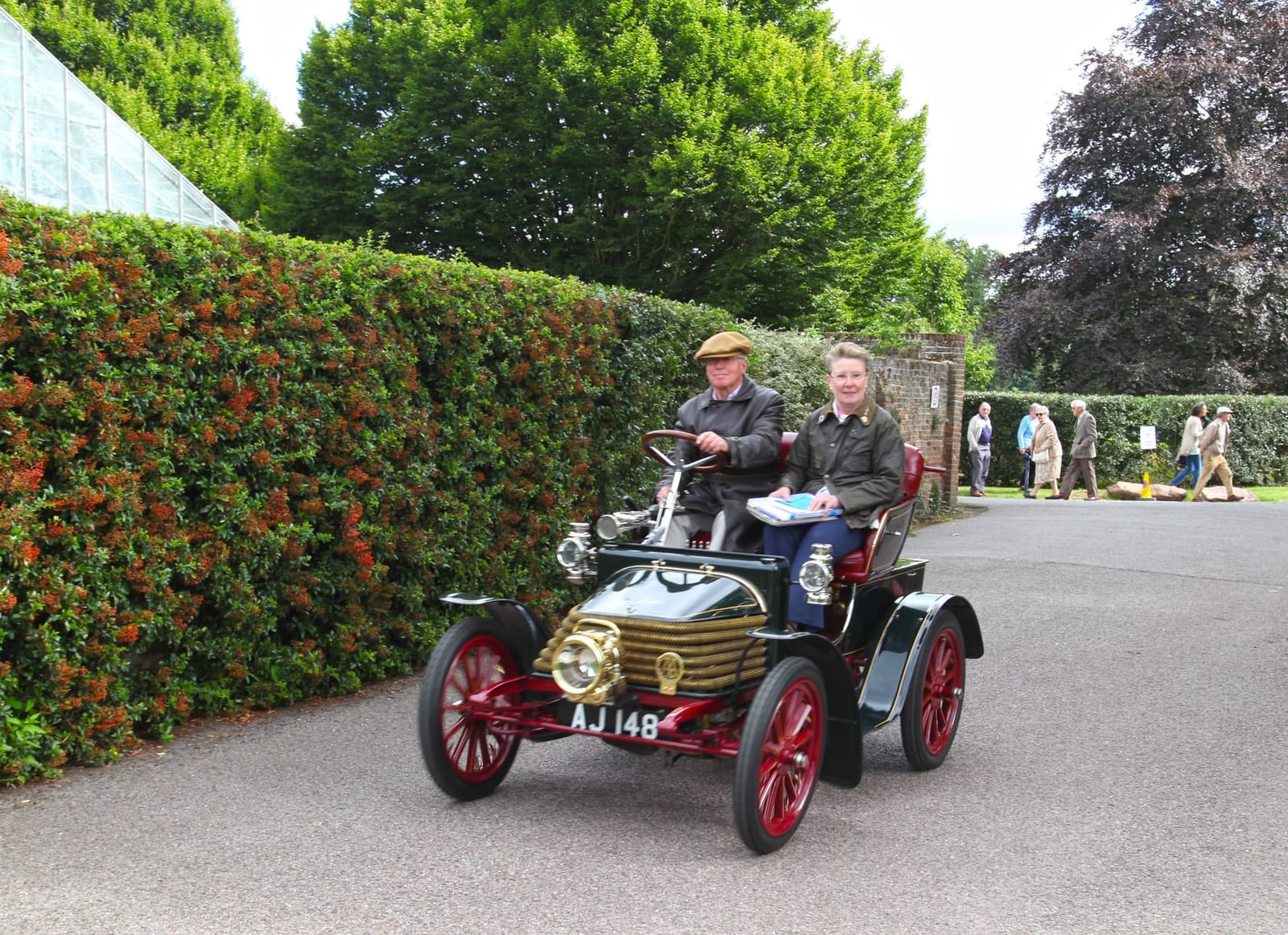 A 1904 Wolseley 6hp veteran car with two occupants driving past a clipped hedge, registration AJ 148, on a run